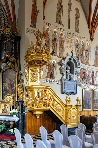 Złocona barokowa ambona zdobiona rzeźbami na tle polichromii w piotrkowskiej świątyni.