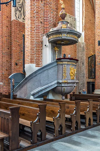 Barokowa ambona z bogatymi złoceniami zamontowana na ceglanym filarze w otoczeniu drewnianych ławek.