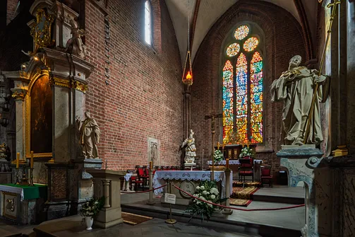 Ceglane ściany prezbiterium świątyni z widocznym ołtarzem, rzeźbami i witrażem.