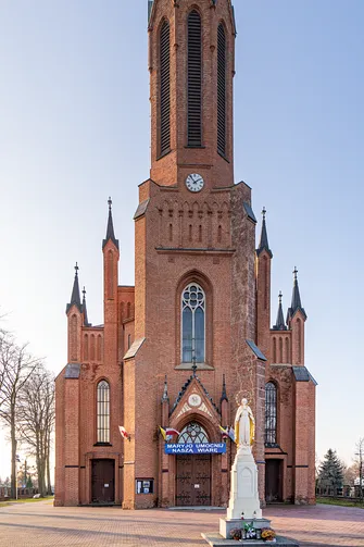 Ceglany neogotycki portal rędzińskiego kościoła z ostrołukowym oknem, zegarem i figurą Maryi.